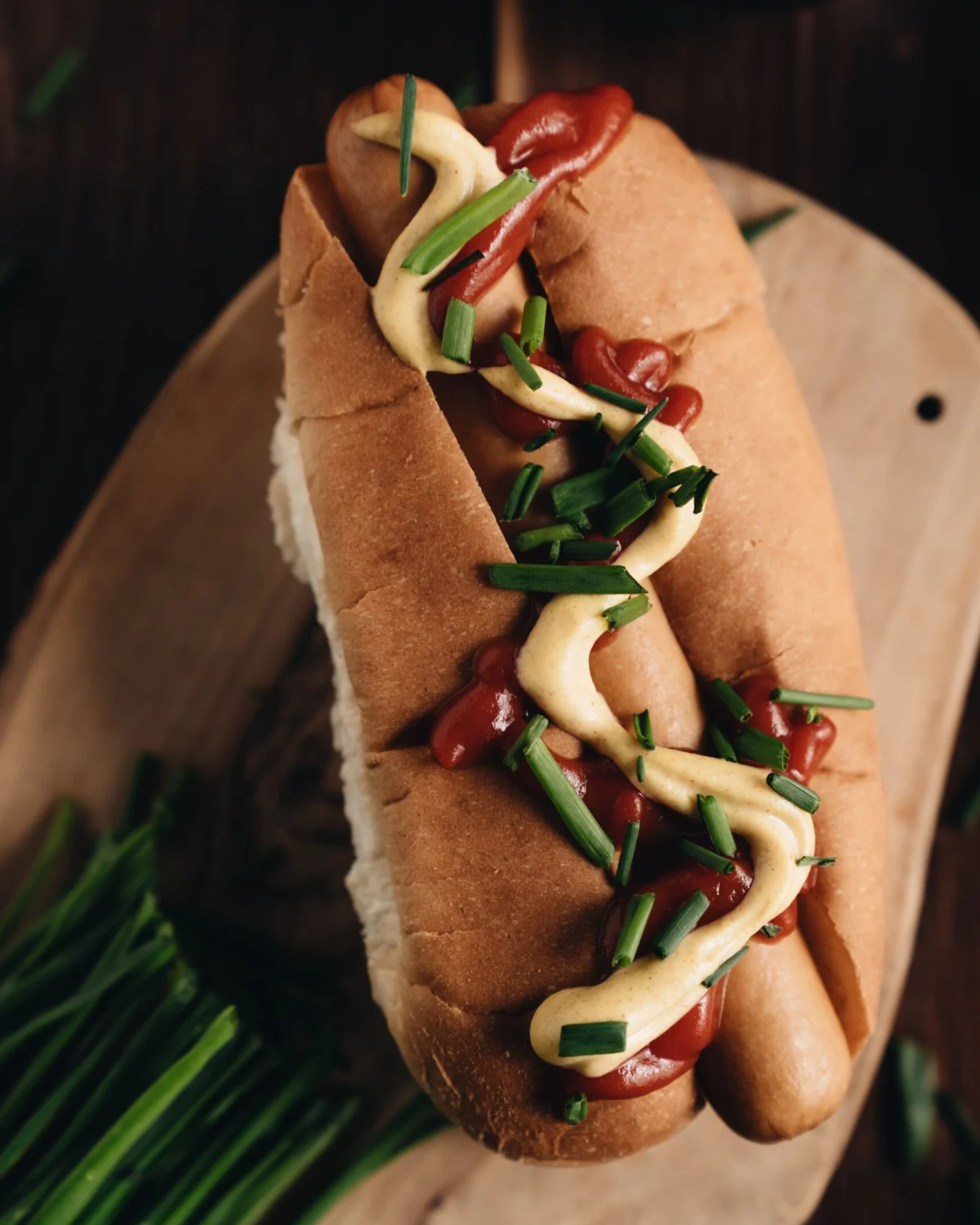 Close-up of a hot dog with mustard, ketchup, and chives on a wooden board